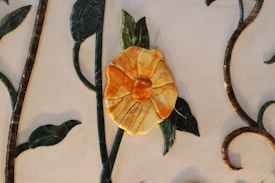 A decorative mosaic artwork featuring a prominent yellow flower with detailed petals set against a backdrop of swirling green leaves and brown curved stems.