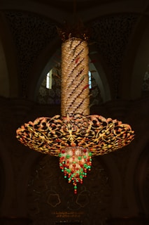 An ornate, oversized chandelier with intricate golden floral patterns is suspended from the ceiling. It features a spiraling column wrapped with lights and decorative elements. The bottom is adorned with dangling crystal-like ornaments in red and green, casting a warm glow. In the background, there's an ornate wall with floral motifs and inscriptions.
