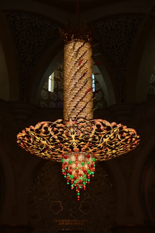 An ornate, oversized chandelier with intricate golden floral patterns is suspended from the ceiling. It features a spiraling column wrapped with lights and decorative elements. The bottom is adorned with dangling crystal-like ornaments in red and green, casting a warm glow. In the background, there's an ornate wall with floral motifs and inscriptions.