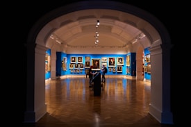 An art gallery features a richly decorated interior with blue walls adorned with classical paintings. The space has a polished wooden floor, and the lighting highlights the artworks. Two individuals are observed near the center, positioned under an archway with a high ceiling, creating a dramatic entrance to the room.