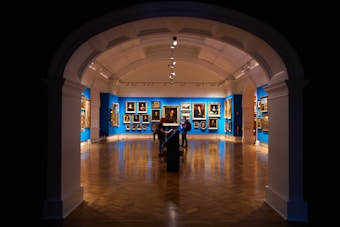An art gallery features a richly decorated interior with blue walls adorned with classical paintings. The space has a polished wooden floor, and the lighting highlights the artworks. Two individuals are observed near the center, positioned under an archway with a high ceiling, creating a dramatic entrance to the room.