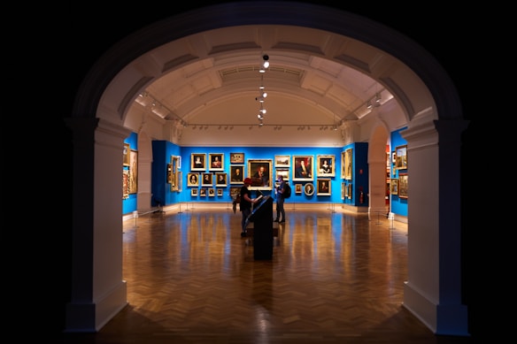 An art gallery features a richly decorated interior with blue walls adorned with classical paintings. The space has a polished wooden floor, and the lighting highlights the artworks. Two individuals are observed near the center, positioned under an archway with a high ceiling, creating a dramatic entrance to the room.