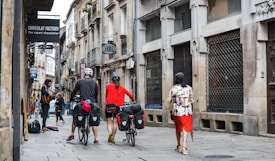 A busy urban street scene features a musician playing a guitar on the left side, while pedestrians walk by. Two cyclists, carrying fully loaded bikes, stand nearby observing the performance. The street is lined with old stone buildings and various shop signs, including one reading 'CHOCOLAT FACTORY.'