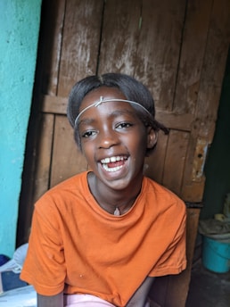 A child wearing an orange shirt smiles cheerfully. The hair is styled neatly with a headband. The background includes a wooden door and a light blue wall, suggesting an indoor setting.