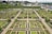 An aerial view of a meticulously landscaped garden displaying symmetrical geometric patterns of various plant beds. The garden is surrounded by hedges and pathways, with a village and church in the background. There are several arches and trellises throughout the garden.