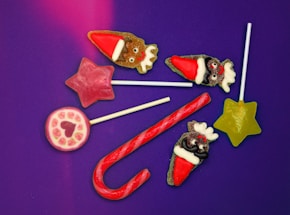 A collection of colorful candies arranged on a purple background. The assortment includes a red and white candy cane, a green star-shaped lollipop, a pink star-shaped lollipop, a round lollipop with rose patterns, and several reindeer-shaped candies wearing Santa hats.