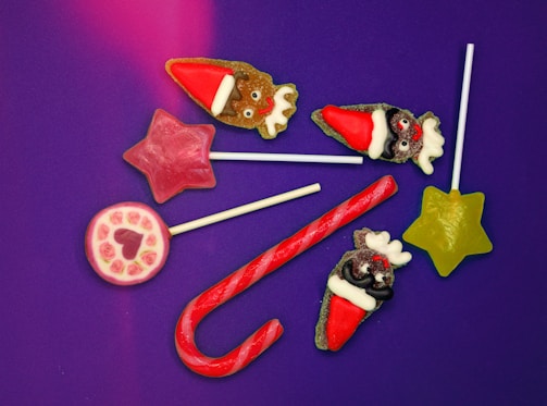 A collection of colorful candies arranged on a purple background. The assortment includes a red and white candy cane, a green star-shaped lollipop, a pink star-shaped lollipop, a round lollipop with rose patterns, and several reindeer-shaped candies wearing Santa hats.