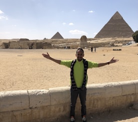A person with outstretched arms stands in front of the Pyramids of Giza and the Great Sphinx in Egypt. The sky is clear with a few clouds, and the ground is sandy. There are other people in the background walking and taking pictures.
