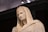 A classical marble statue depicting a serene figure draped in a cloak with a hood. The figure has a calm and thoughtful expression, and the craftsmanship highlights intricate details on the face and garments.