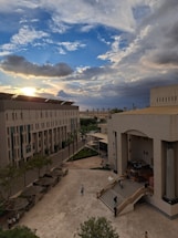 A university campus scene with a wide courtyard is surrounded by modern architectural buildings, featuring steps and outdoor seating areas. The scene captures a vibrant sky filled with dramatic clouds and the sun peeking through, casting a warm glow over the scene. Several people are visible walking through the courtyard.