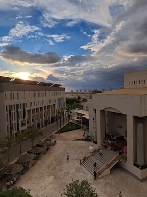 A university campus scene with a wide courtyard is surrounded by modern architectural buildings, featuring steps and outdoor seating areas. The scene captures a vibrant sky filled with dramatic clouds and the sun peeking through, casting a warm glow over the scene. Several people are visible walking through the courtyard.