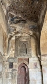 A richly detailed stone wall is adorned with intricate carvings and inscriptions. The arch design frames a section with abstract patterns and scripts, possibly of historical or religious significance. The stonework features varying shades and textures, contributing to a rustic yet majestic appearance.
