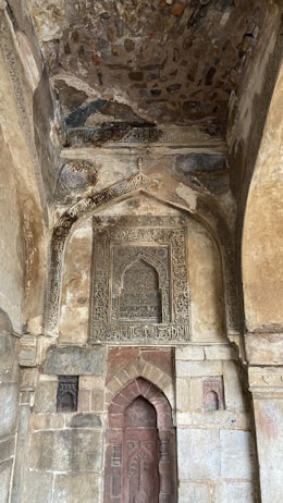A richly detailed stone wall is adorned with intricate carvings and inscriptions. The arch design frames a section with abstract patterns and scripts, possibly of historical or religious significance. The stonework features varying shades and textures, contributing to a rustic yet majestic appearance.