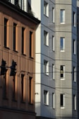 A modern urban setting with two adjacent buildings, one in a warm brown tone and the other in a light gray. The structures feature multiple windows and intersecting power lines. The scene is highlighted by shadows creating contrasting patterns on the building surfaces.