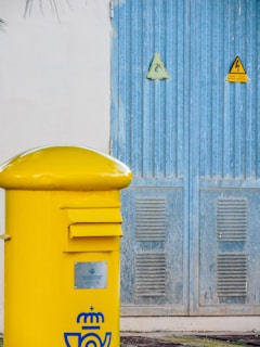A bright yellow postal box is situated in the foreground, marked with a symbol indicative of postal services. Behind it, a large blue metal door is visible, featuring warning signs with electrical hazard symbols.