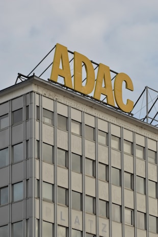 A modern building facade with a grid-like pattern of windows, featuring a prominent sign on the rooftop displaying yellow letters ADAC against a partly cloudy sky.