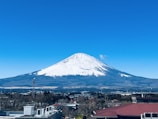 A snow-capped mountain with a pointed peak dominates the background, under a clear blue sky. In the foreground, a town with various buildings and structures is visible, nestled at the base of the mountain.