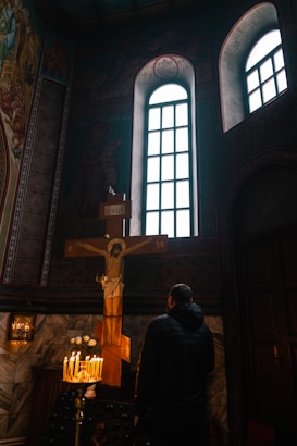 A person is standing in a dimly lit church, facing a large cross with a depiction of Jesus on it. The scene is illuminated by a cluster of lit candles on a stand below the cross, casting a warm glow. The background features intricately decorated walls and tall, narrow windows allowing light to filter in.
