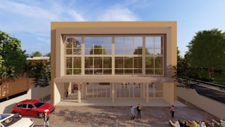 A modern, two-story building with a large glass facade, surrounded by greenery. People are visible near the entrance and a red car is parked in front.