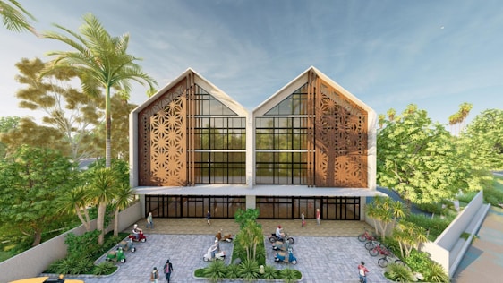 A modern two-story building with an ornate facade featuring geometric patterns and large windows. The structure is surrounded by lush green trees and palm trees, enhancing the natural setting. In front of the building, there is a paved courtyard with several people walking and bicycles parked. The sky is clear and blue, adding to the serene environment.
