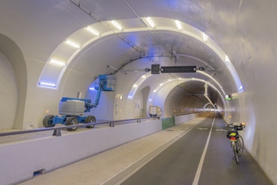 A well-lit tunnel with white and blue lighting features a long walkway. On the left side, there is a blue elevated work platform, while on the right side, a bicycle is parked. The tunnel is spacious with smooth walls and is equipped with various signs and safety barriers.