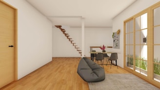 A modern, minimalistic interior features a wooden floor and white walls. Wooden stairs without railings ascend to an upper level in the back corner. A simple dining table with a pair of gray chairs is positioned near a wall decorated with a grid and a plant. A soft, gray bean bag rests on a light gray rug, near a large wooden-framed window that lets in natural light.