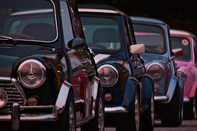 Classic cars are parked in a row, featuring shiny, reflective surfaces and distinct round headlights. The cars are lined up closely together, showcasing a variety of bold, vibrant colors against a dark background.