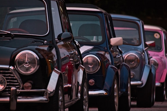Classic cars are parked in a row, featuring shiny, reflective surfaces and distinct round headlights. The cars are lined up closely together, showcasing a variety of bold, vibrant colors against a dark background.