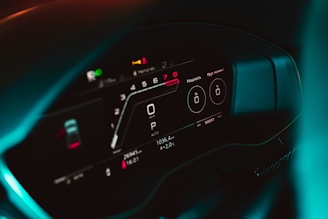 A close-up of a car's digital dashboard provides detailed instrument readings, including speed, gear position, and various indicators. The display features a sleek, modern design with a dark background and illuminated white and red text. The overall look is futuristic and minimalistic.
