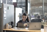 A woman is sitting at a desk, reading a book or magazine. There are office supplies like a pen holder and mug on the desk, as well as a laptop. The workspace is partitioned by a soft gray divider with pinned notes or photos. The background shows a modern office environment with people blurred in the distance.