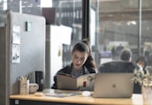 A woman is sitting at a desk, reading a book or magazine. There are office supplies like a pen holder and mug on the desk, as well as a laptop. The workspace is partitioned by a soft gray divider with pinned notes or photos. The background shows a modern office environment with people blurred in the distance.