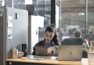 A woman is sitting at a desk, reading a book or magazine. There are office supplies like a pen holder and mug on the desk, as well as a laptop. The workspace is partitioned by a soft gray divider with pinned notes or photos. The background shows a modern office environment with people blurred in the distance.