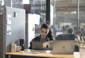 A woman is sitting at a desk, reading a book or magazine. There are office supplies like a pen holder and mug on the desk, as well as a laptop. The workspace is partitioned by a soft gray divider with pinned notes or photos. The background shows a modern office environment with people blurred in the distance.