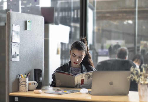 A woman is sitting at a desk, reading a book or magazine. There are office supplies like a pen holder and mug on the desk, as well as a laptop. The workspace is partitioned by a soft gray divider with pinned notes or photos. The background shows a modern office environment with people blurred in the distance.