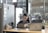 A woman is sitting at a desk, reading a book or magazine. There are office supplies like a pen holder and mug on the desk, as well as a laptop. The workspace is partitioned by a soft gray divider with pinned notes or photos. The background shows a modern office environment with people blurred in the distance.