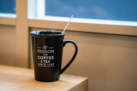 A black mug with white text sits on a wooden surface. The words 'Passion for Coffee & Tea Since 1963' are printed on the mug. A wooden stir stick leans inside the mug. The background shows a window with white frames.