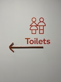 A minimalist toilet sign with stylized male and female figures in orange, accompanied by the word 'Toilets' and an arrow pointing to the left.