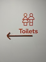 A minimalist toilet sign with stylized male and female figures in orange, accompanied by the word 'Toilets' and an arrow pointing to the left.