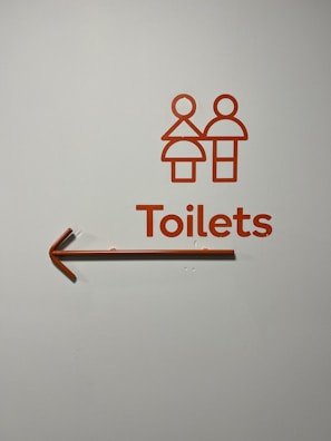 A minimalist toilet sign with stylized male and female figures in orange, accompanied by the word 'Toilets' and an arrow pointing to the left.