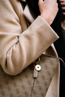 A close-up view of a person wearing a beige coat, holding a designer handbag with a recognizable pattern and a keychain that has the brand logo and name attached. The person is also wearing a black garment underneath.
