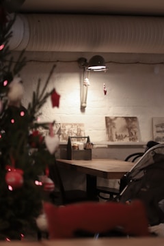 A cozy, dimly lit interior featuring a wooden table with chairs, a decorated Christmas tree, and a wall-mounted light. The setting includes a stroller and artistic wall decorations, creating a warm and inviting atmosphere.