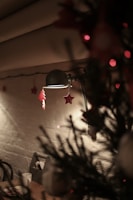 A cozy indoor setting with soft lighting. A vintage lamp casts a warm glow, highlighting festive ornaments such as stars and trees hanging from its arm. In the foreground, blurred branches of a Christmas tree add a seasonal touch. A small table with cups and plates is visible in the background against a textured wall.