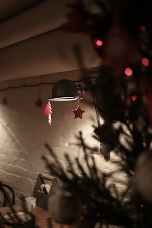 A cozy indoor setting with soft lighting. A vintage lamp casts a warm glow, highlighting festive ornaments such as stars and trees hanging from its arm. In the foreground, blurred branches of a Christmas tree add a seasonal touch. A small table with cups and plates is visible in the background against a textured wall.