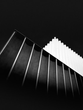 A black and white photograph featuring modern architectural lines and shapes. The building includes a series of vertical slats, showing a contrast between light and shadow. The image captures a sense of geometric abstraction with an emphasis on texture and form.