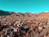 A town is situated in a valley surrounded by mountainous terrain. There are numerous buildings and structures with a mix of greenery and barren land. The mountains in the background have varying shades of brown and some snow-capped peaks.