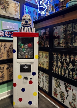 A vintage clown-themed vending machine stands in the foreground, adorned with colorful polka dots and featuring a clown head on top with a red nose and blue eyes. The machine is surrounded by framed tattoo flash art, displaying a variety of traditional tattoo designs including animals, pin-up girls, and other classic motifs. A neon bunny sign glows in blue and white in the upper right corner, adding to the eclectic ambiance of the room.