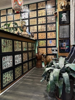 The room features a wall adorned with an extensive display of framed tattoo flash art showcasing diverse designs. A variety of trinkets and sculptures, including a bust and animal statues, are placed around the space. Several wall hangings, including t-shirts and a humorous 'Funeral No Parking' sign, add to the eclectic decor. A large plant occupies the foreground, contributing to the room's organic feel. The ambiance is enhanced by wood flooring and a ceiling adorned with a decorative lantern.