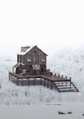A wooden house stands on stilts surrounded by a snowy landscape, with snow-covered trees in the background. The architecture is rustic, with metal roofing and a sloped walkway leading to the entrance.