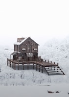 A wooden house stands on stilts surrounded by a snowy landscape, with snow-covered trees in the background. The architecture is rustic, with metal roofing and a sloped walkway leading to the entrance.