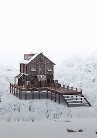 A wooden house stands on stilts surrounded by a snowy landscape, with snow-covered trees in the background. The architecture is rustic, with metal roofing and a sloped walkway leading to the entrance.